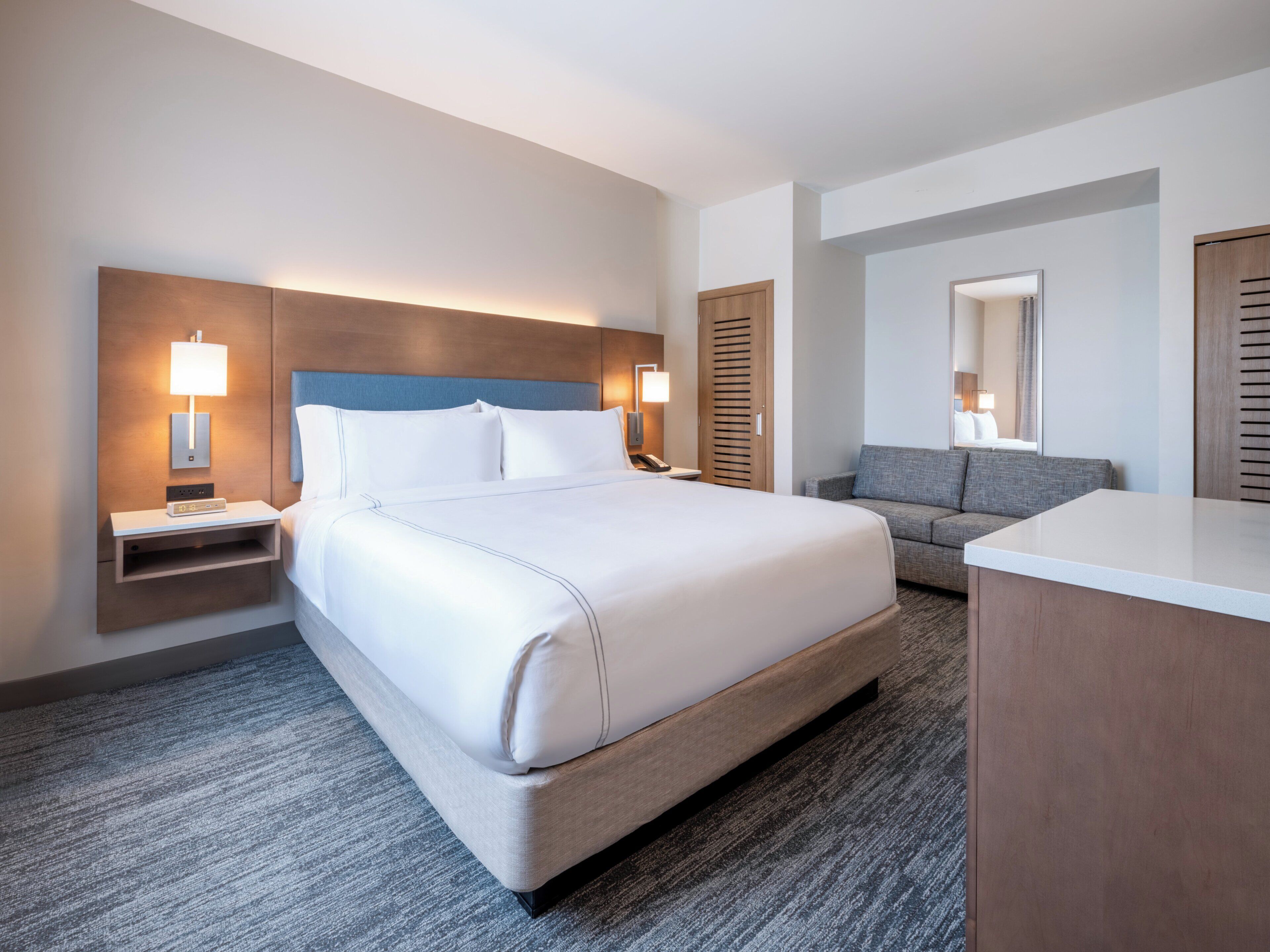 A hotel room with a large bed, bedside tables with lamps, a sofa, a desk, and a mirror.