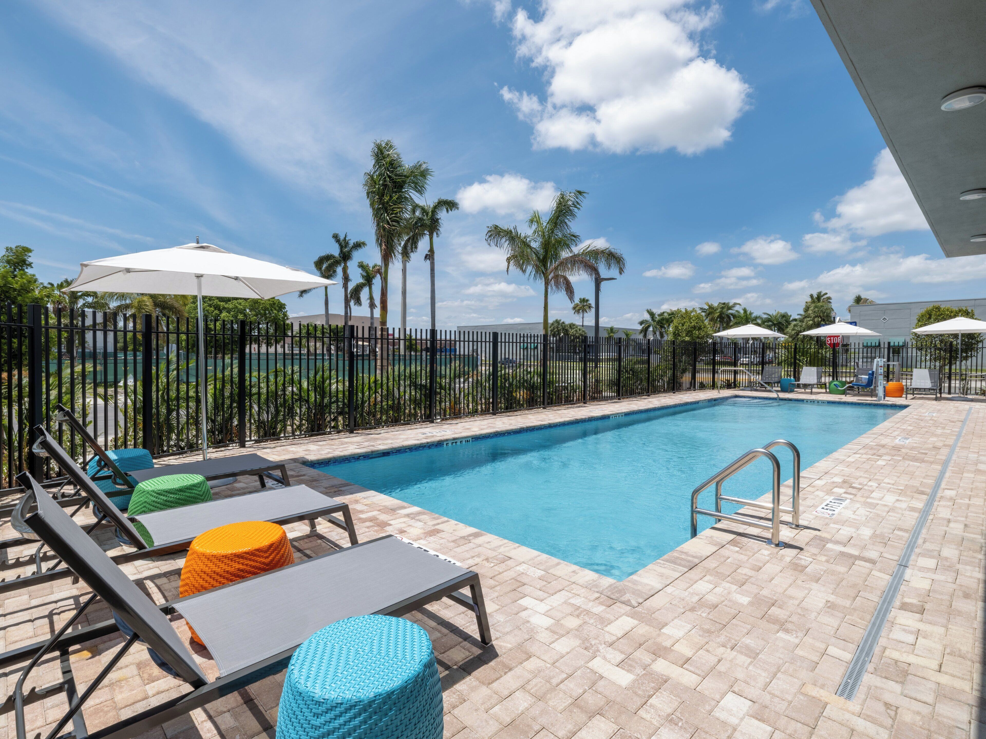 A swimming pool with lounge chairs and umbrellas, surrounded by palm trees and a fence.
