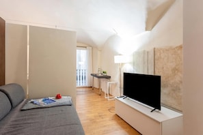 Un salon moderne avec un canapé gris, un téléviseur à écran plat, une petite table avec une lampe et une vue sur un balcon.