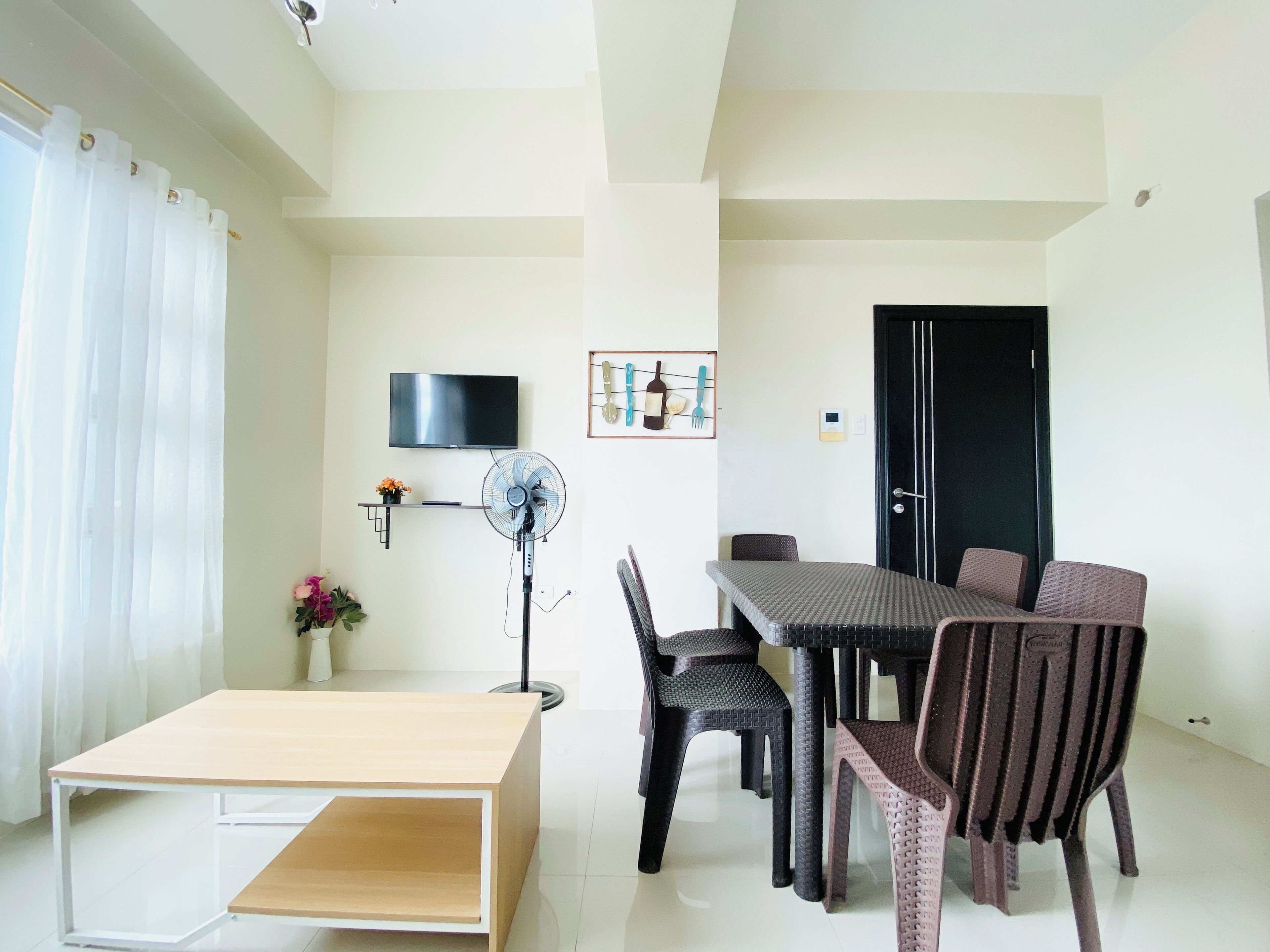 A modern living room with a flat-screen TV, a standing fan, a dining table with chairs, and a small side table.