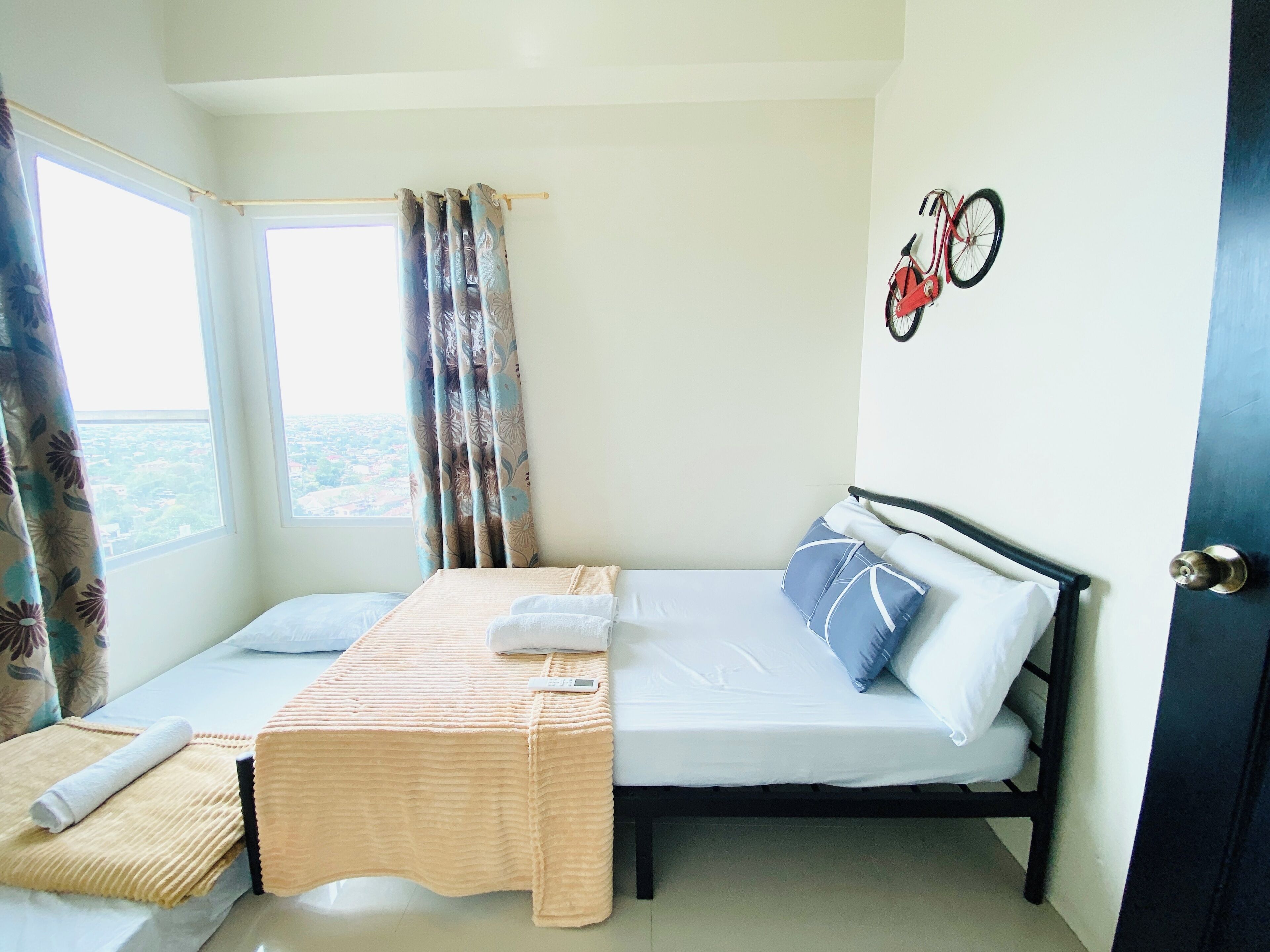 A small, well-lit room with a bed, a bedside table, a window with curtains, and a bicycle mounted on the wall.