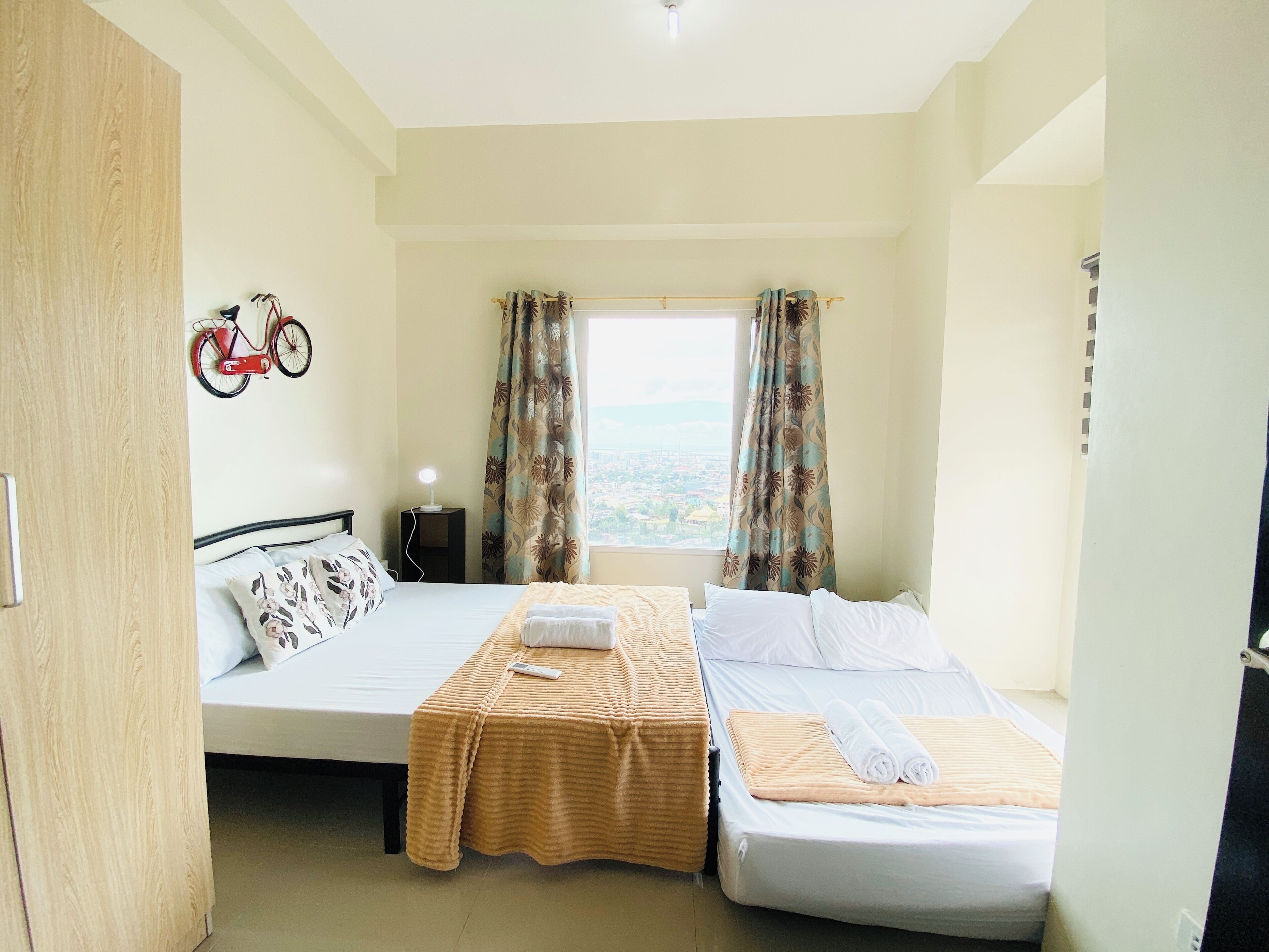 Una habitación con dos camas, una mesita de noche, una lámpara, una decoración con motivo de bicicleta y una ventana con cortinas.