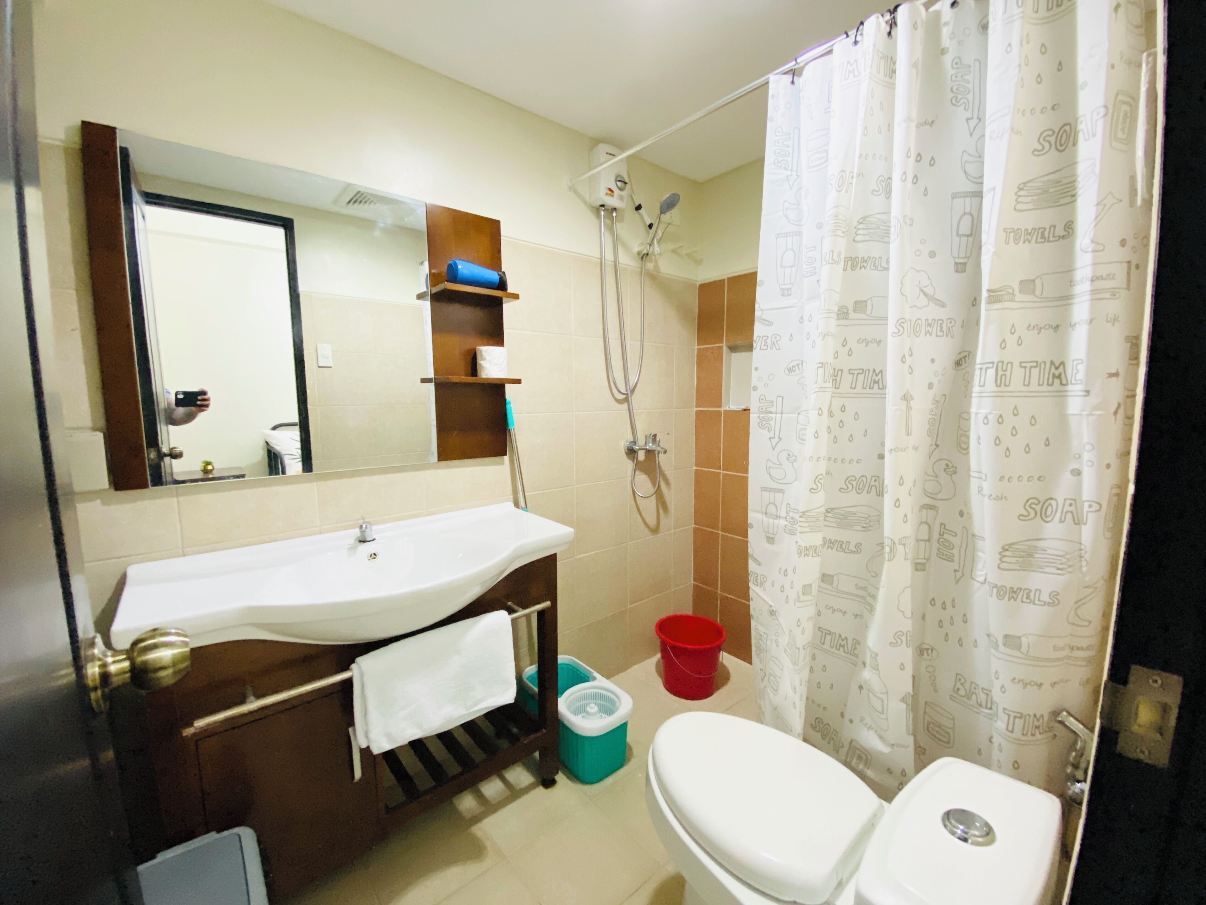 A bathroom with a white sink, mirror, and shower. There is a towel rack and a red bucket on the floor.