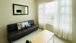 Ein Wohnzimmer mit einem dunklen Sofa, einem Holztisch, einem großen Fenster mit transparenten Vorhängen und einer Chandelüster.