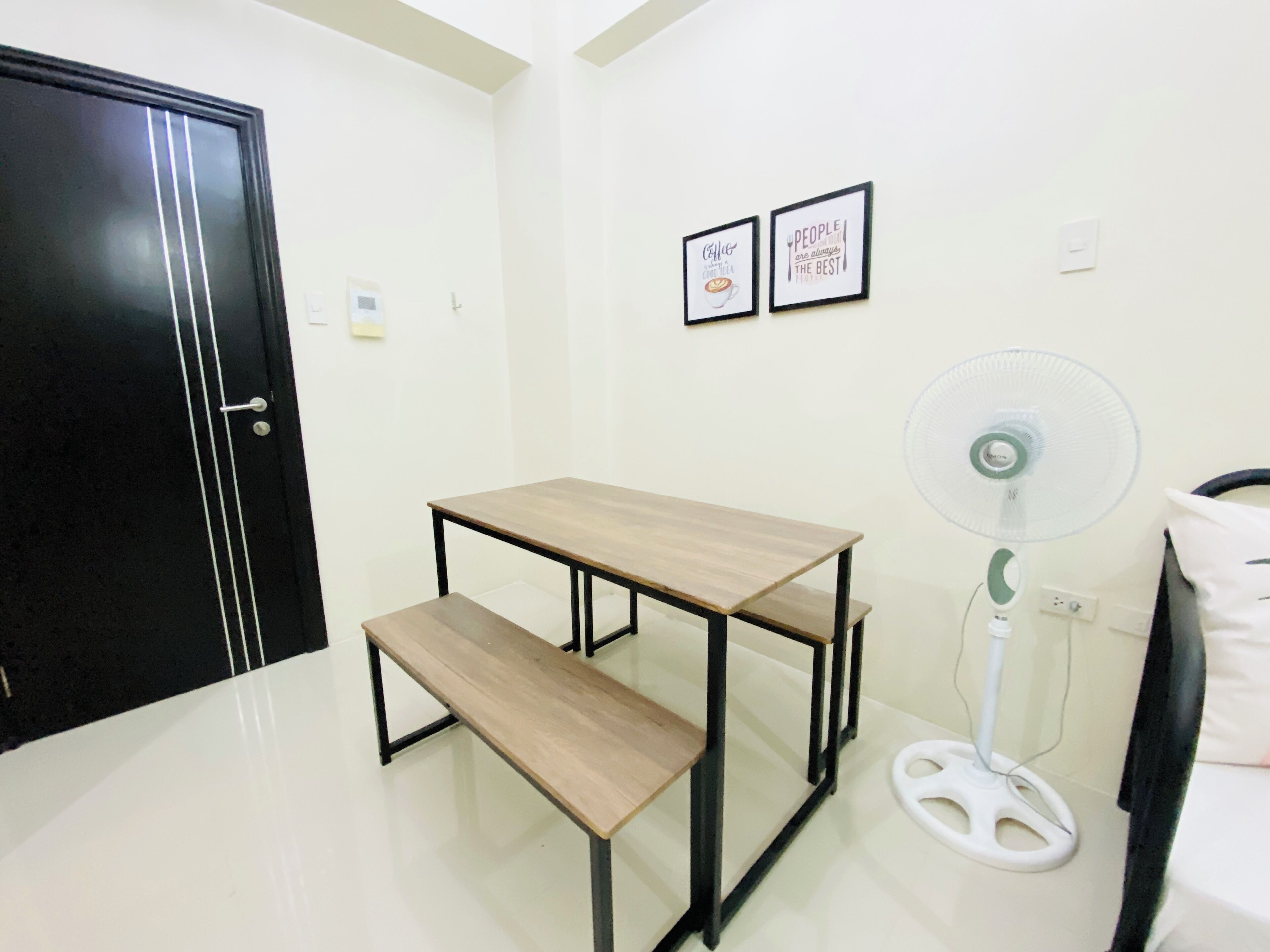 Una habitación pequeña con una mesa y un banco de madera, un ventilador blanco y dos cuadros en la pared.