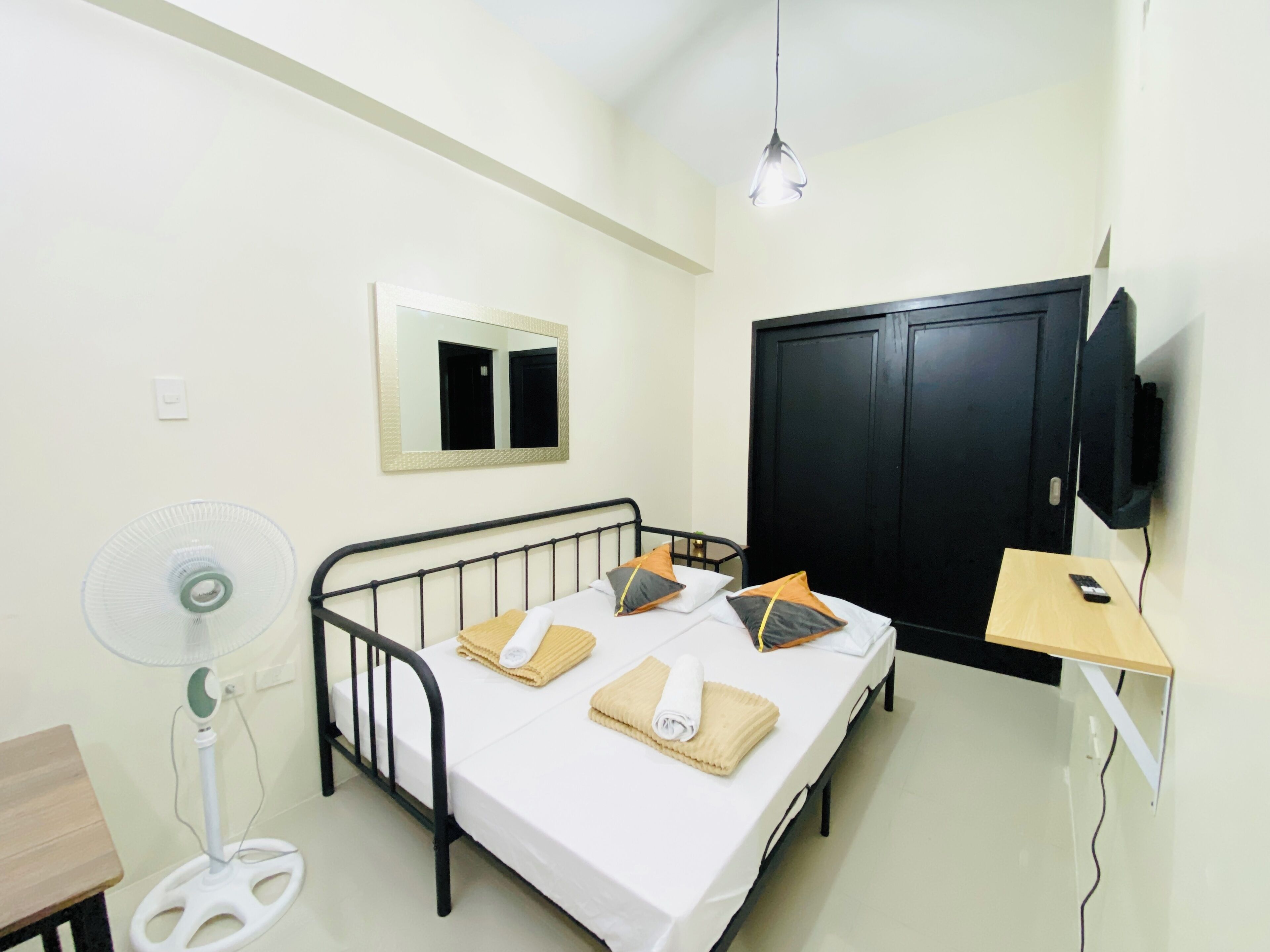 Una cama individual con almohadas, una mesita de noche, un televisor, un ventilador y un armario en una habitación con paredes blancas.