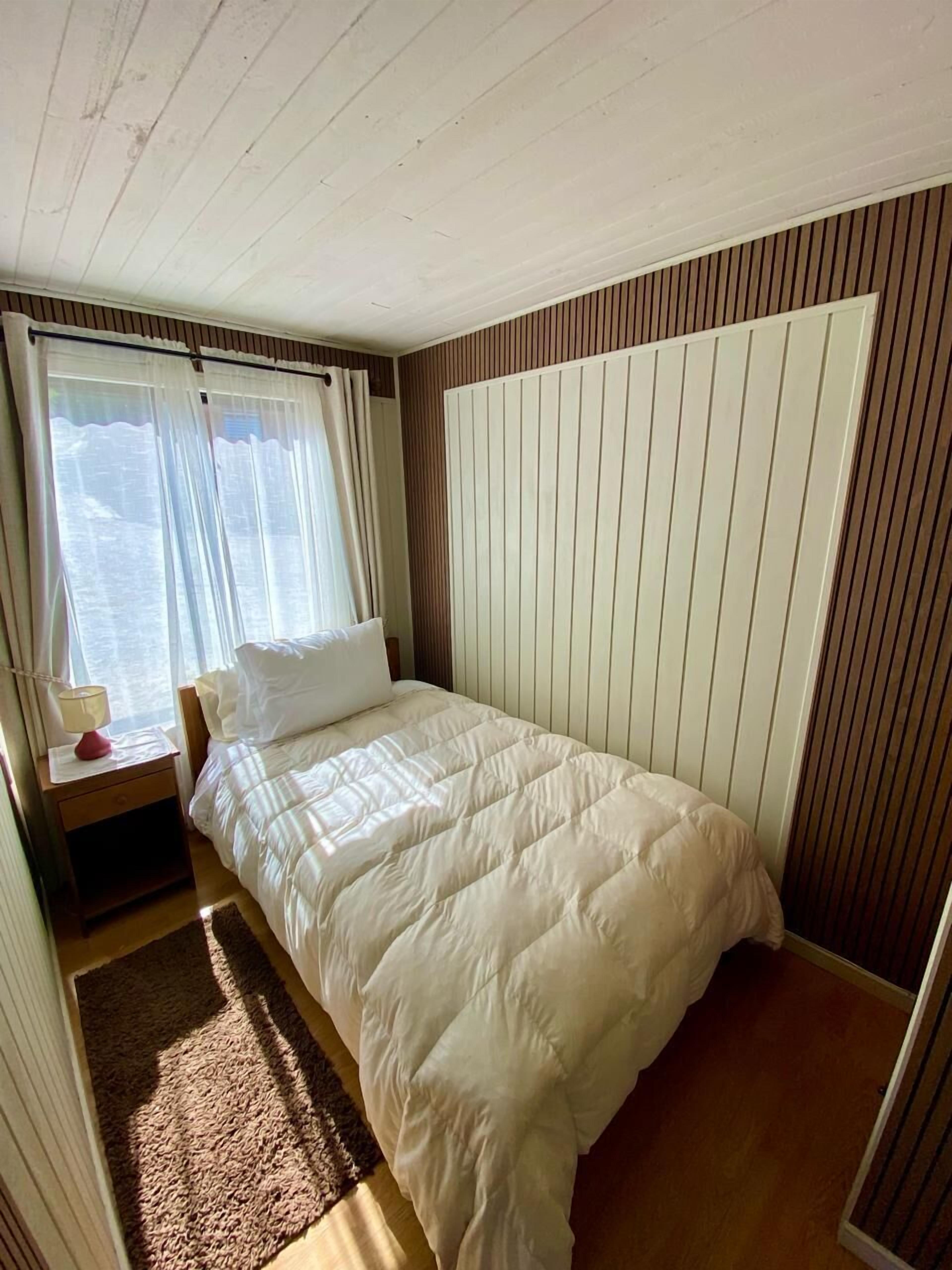 Una camera da letto con un letto, un comodino, una finestra con tende e una parete rivestita in legno.