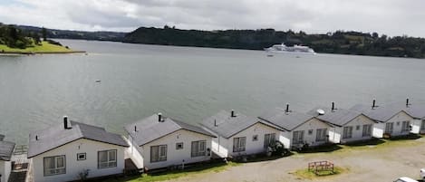 A row of single-story cabins with a view of a lake and a cruise ship in the distance.