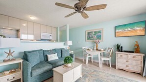 A living room with a teal sofa, a white coffee table, a ceiling fan, a kitchen area with white cabinets, and a dining table with chairs.