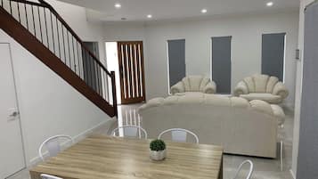 En moderne stue med et træspisebord, hvide stole, en beige sofa og en trappe.