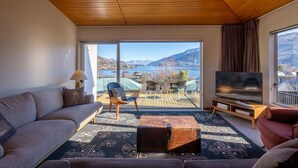 Una sala de estar con un sofá, una mesa de centro de madera y un televisor. Hay un ventanal con vistas a un lago y montañas.