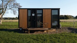 Una cabaña moderna de un solo piso con grandes puertas y ventanales de vidrio, una terraza de madera y una pequeña zona de estar al aire libre.