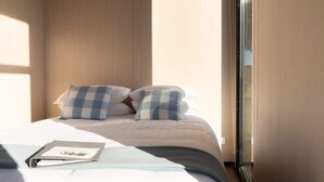 A neatly made bed with checkered pillows, a bedside table with a book, and a window with blinds.