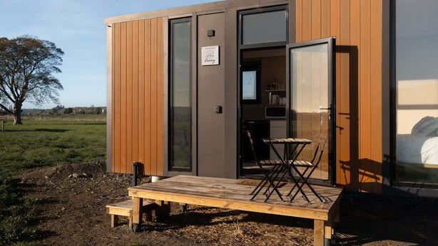 A modern cabin with a wooden exterior, a sliding glass door entrance, a small dining area with a table and chairs, and a bench outside.