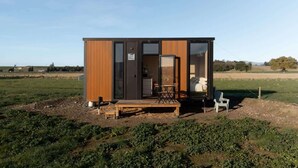A small, modern cabin with a wooden exterior and a glass door leading to an interior with a bed, a table, and a chair.