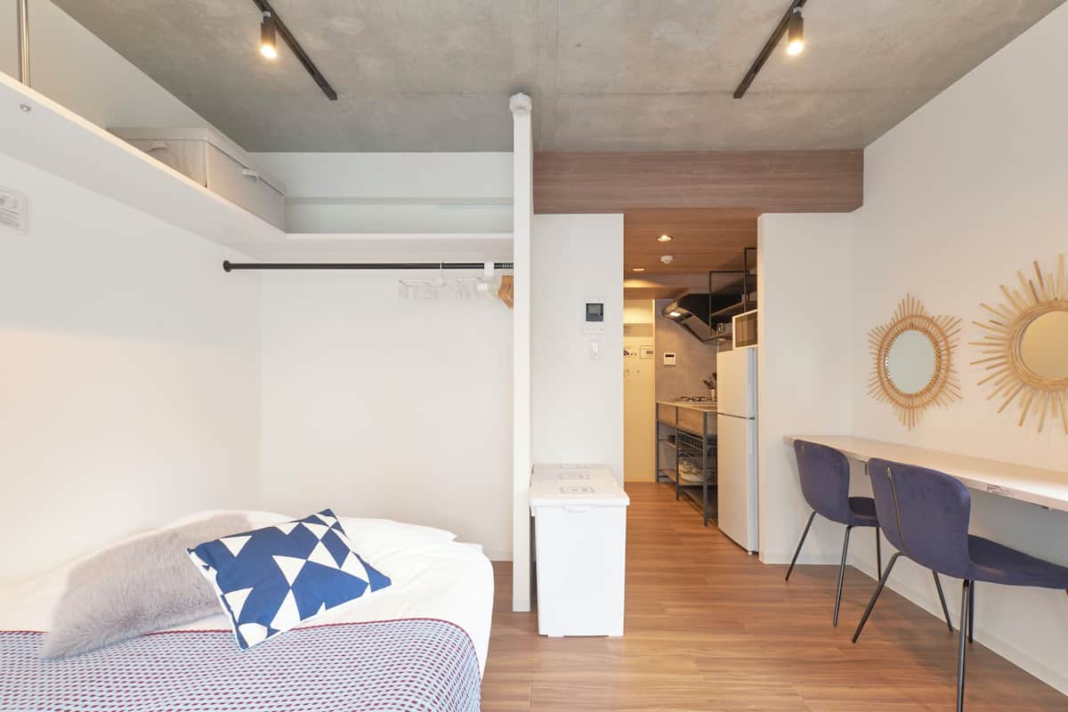Quarto de hotel compacto com cama, uma pequena escrivaninha e uma cadeira. Uma kitchenette é visível ao fundo.