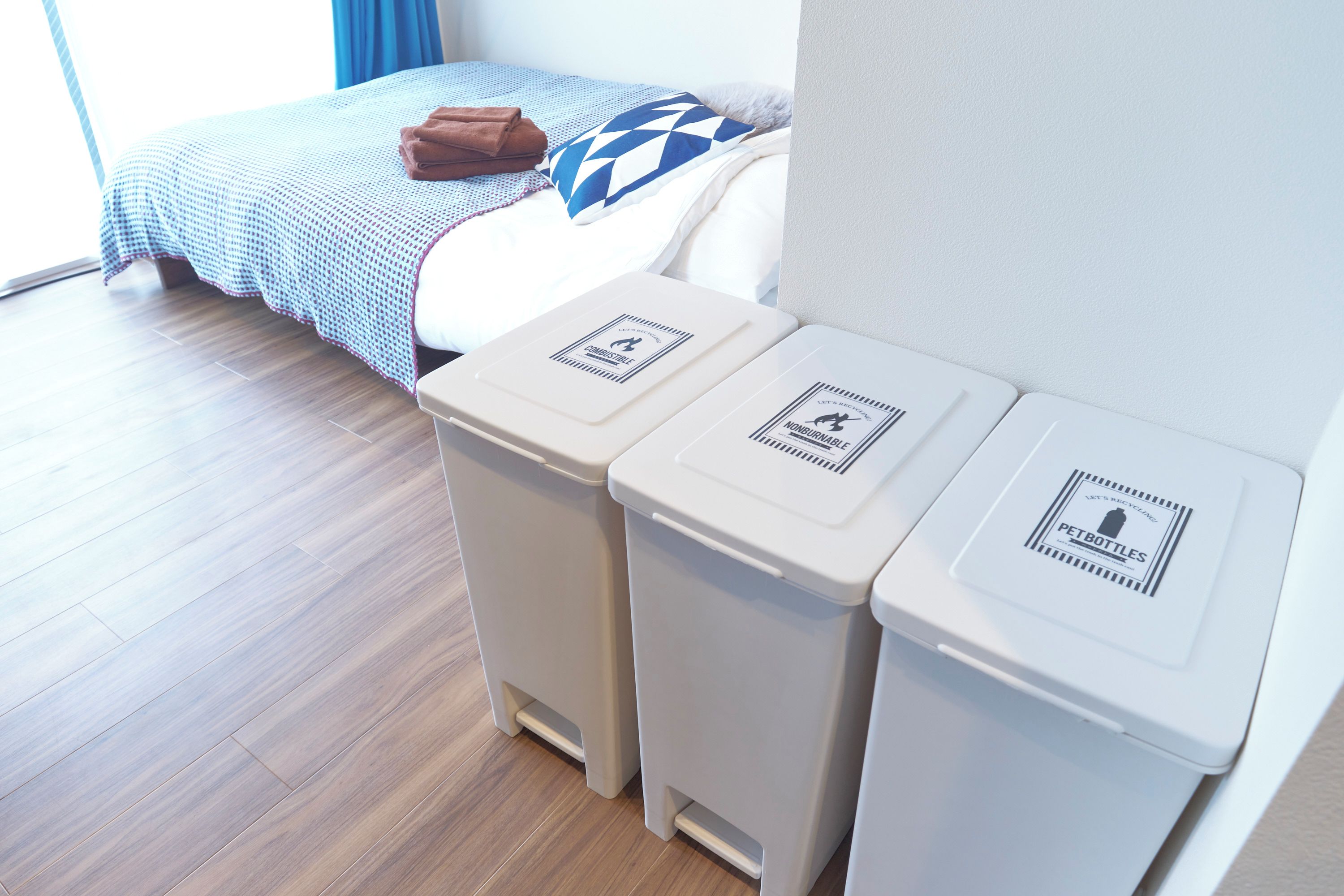 Hotelkamer met een bed, een blauw-wit beddekleed, een bruine handdoek en twee afvalbakken met recyclinglabels.