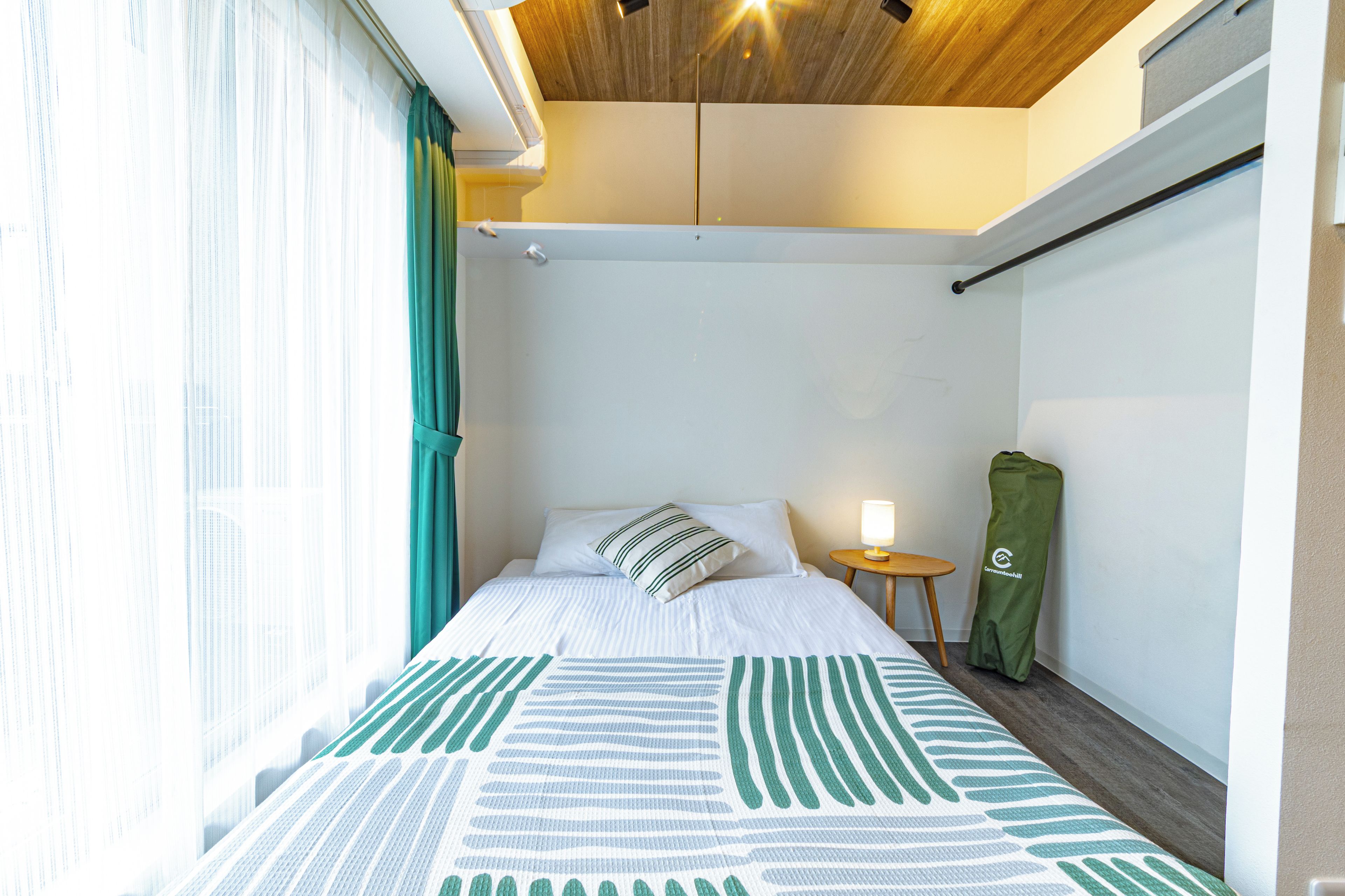 Una habitación de hotel con cama, una mesita de noche con lámpara, un bolso verde y una ventana con cortinas.