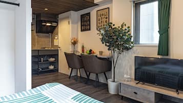 A modern living room with a flat-screen TV, a dining area with chairs and a table, and a kitchen with a stove and cabinets.