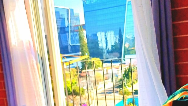 A bedroom with a bed, a window with curtains, and a view of buildings and greenery outside.