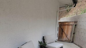 Uma área de pátio com duas cadeiras cinza acolchoadas e uma pequena mesa, um tapete tecido e um portão de madeira ao fundo.