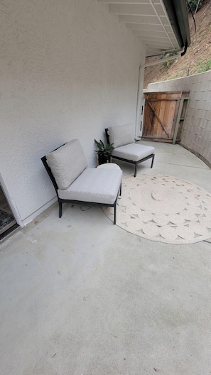 Un espace patio avec deux fauteuils gris rembourrés et une petite table, un tapis tissé et, au fond, un portail en bois.