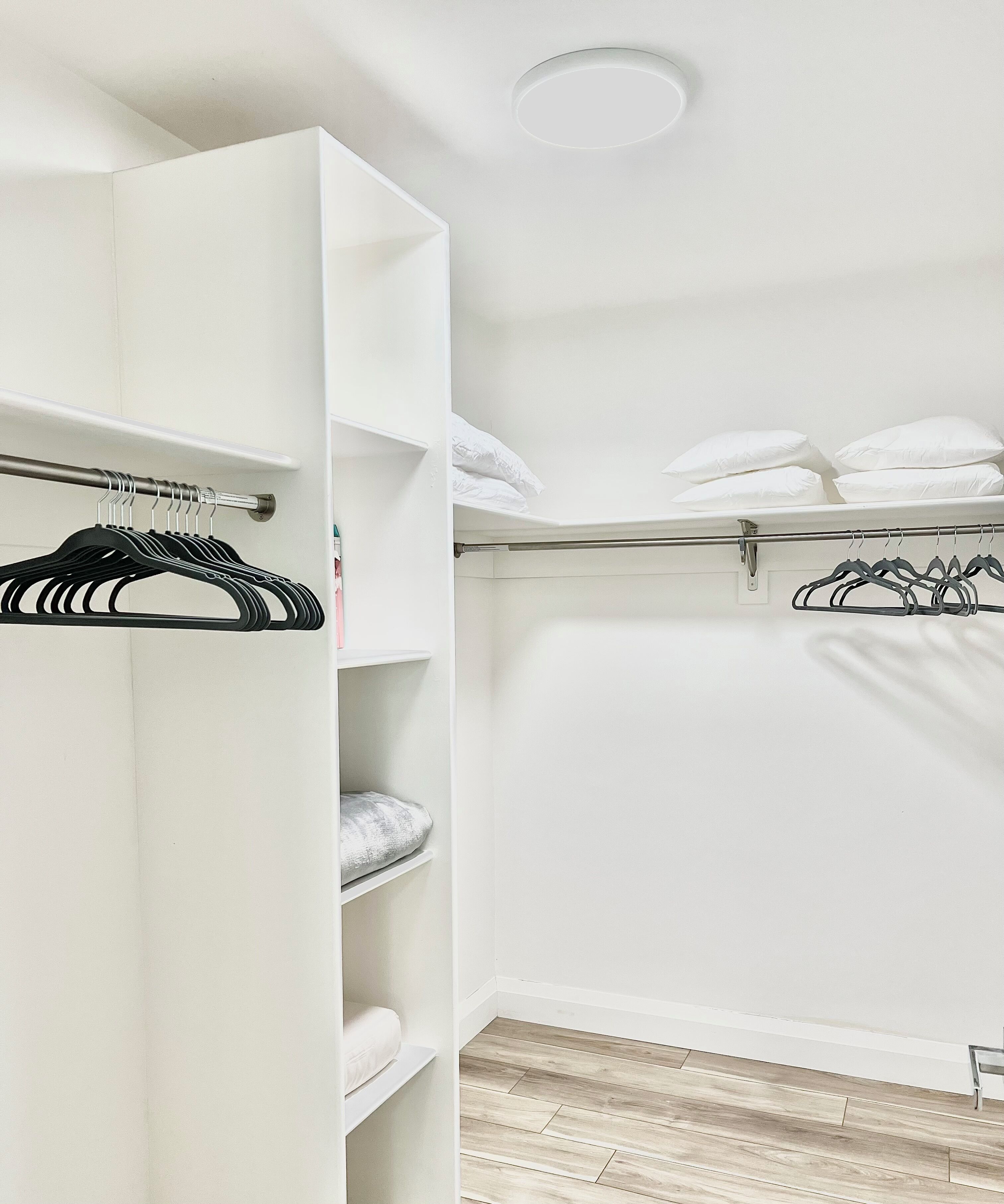 A white walk-in closet with shelves and hanging space, a wooden floor, and a ceiling light.