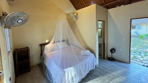 Un dormitorio con una cama bajo una mosquitera, un ventilador de pie y vista a un jardín desde una puerta abierta.