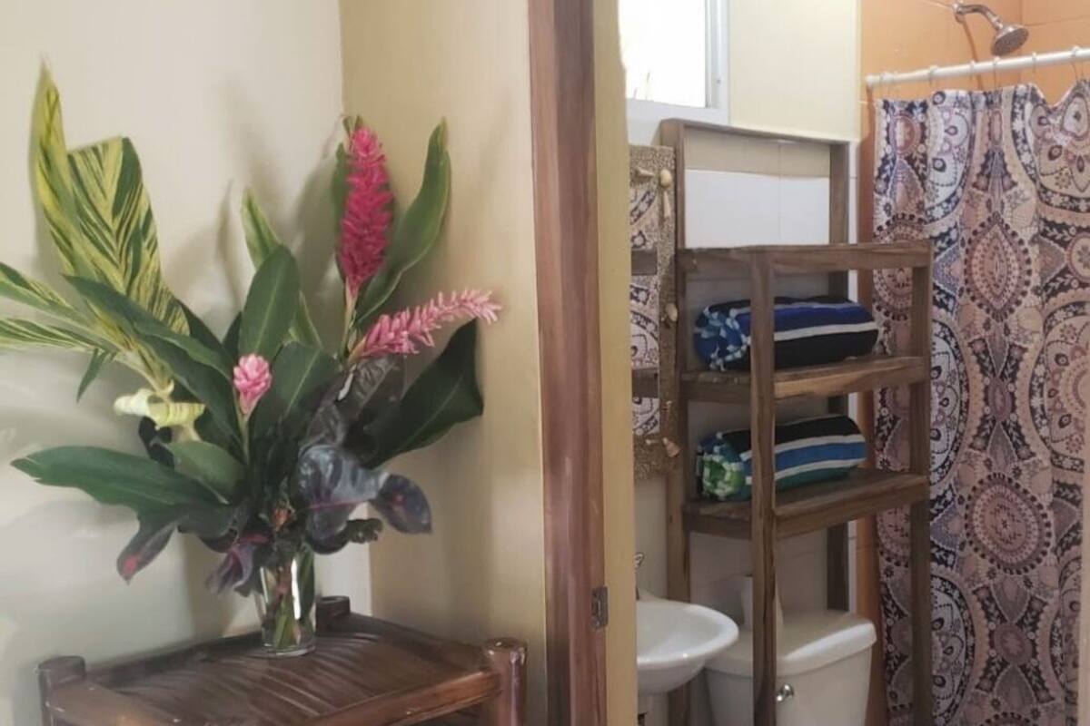A bathroom with a toilet, sink, and shower area. There is a wooden shelf with a vase of flowers and a small table.