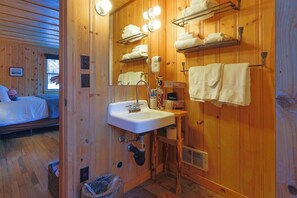 Bagno di una baita in legno, con lavandino, specchio, portasci e un letto visibile nella stanza adiacente.