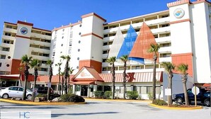 Un hôtel à étages, avec une entrée principale surmontée d’un toit rouge et des palmiers en premier plan.