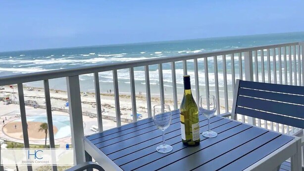 Un balcon avec vue sur l’océan et la plage, une bouteille de vin et deux verres posés sur une table.