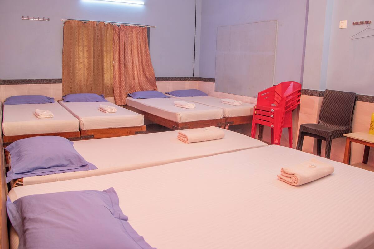 A hotel room with four single beds, each with a pillow and a folded towel. There are curtains on the windows, a small table with a chair, and a TV on a stand.