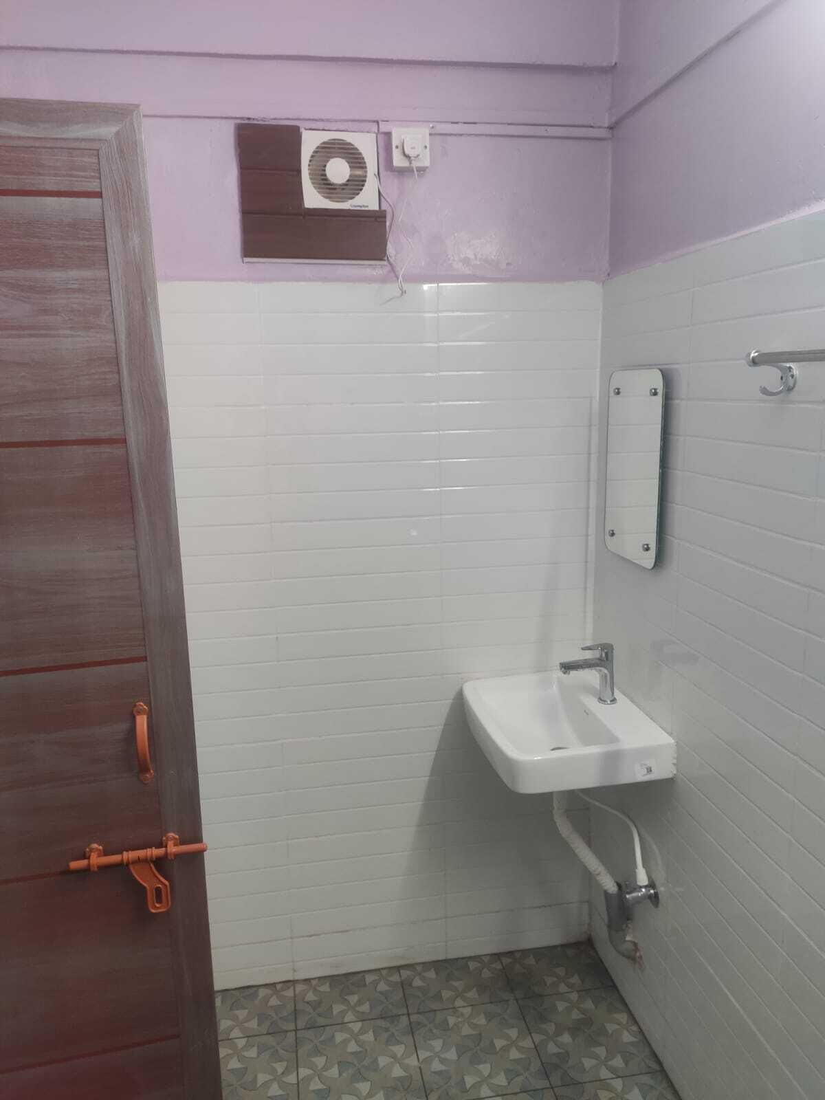 Un baño pequeño con paredes de azulejos blancos, una puerta de madera, un lavamanos blanco y una unidad de ventilación sobre el lavamanos.