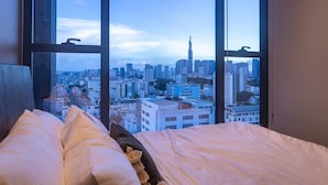 A bed with a cityscape view through large windows, a wooden headboard, and a fluffy pillow. - The MARQ  By An Gia Hospitality (Ho Chi Minh City)