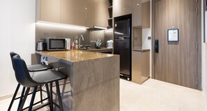 A modern kitchen with a central island, bar stools, a microwave, a refrigerator, and a wooden door. - The MARQ  By An Gia Hospitality (Ho Chi Minh City)