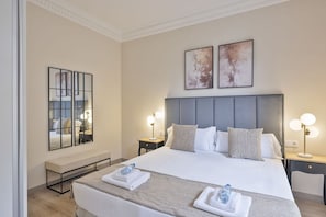 Quarto de hotel com uma cama grande, duas mesas de cabeceira, um banco e obras de arte nas paredes.