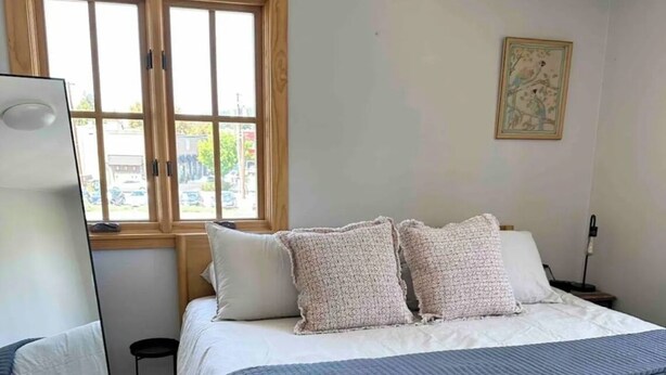 Ein ordentlich bezogenes Bett mit weißer und blauer Bettwäsche, ein hölzerner Kopfteil und ein Fenster mit Blick auf die Straße.