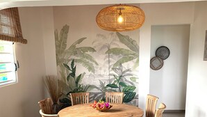 A dining area with a round wooden table surrounded by six chairs, a woven light fixture above, and a mural of tropical foliage on the wall.