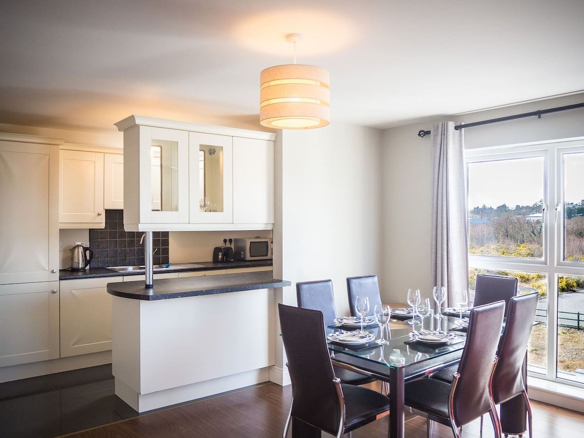 Cozinha moderna com armários brancos, uma ilha central e um espaço de jantar com mesa e cadeiras de vidro.