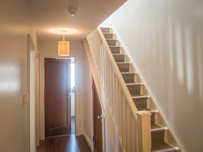 A hallway with a wooden door at the end, a staircase with a white railing, and a hanging light fixture.