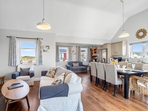 Un vaste espace de vie comprenant un canapé, une table à manger et des chaises, une cuisine équipée de placards et d’un réfrigérateur, ainsi que de grandes fenêtres offrant une vue dégagée.