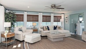 A living room with a sofa, ottoman, and coffee table, a ceiling fan, and a large window with blinds.