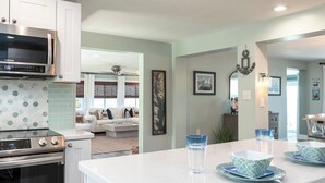 A modern kitchen with white cabinets, a stainless steel microwave, and a built-in oven. There is a dining area with a white table and blue patterned bowls.