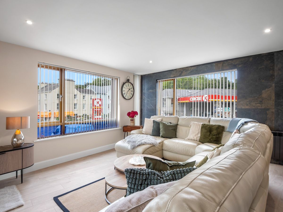 A modern living room with a large sofa, a coffee table, and a side table. There is a window with a view of buildings and a clock on the wall.