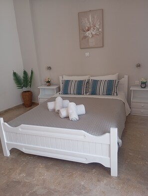 A bedroom with a white bed, a bedside table, a nightstand, a wall-mounted light, a framed artwork, and a potted plant.