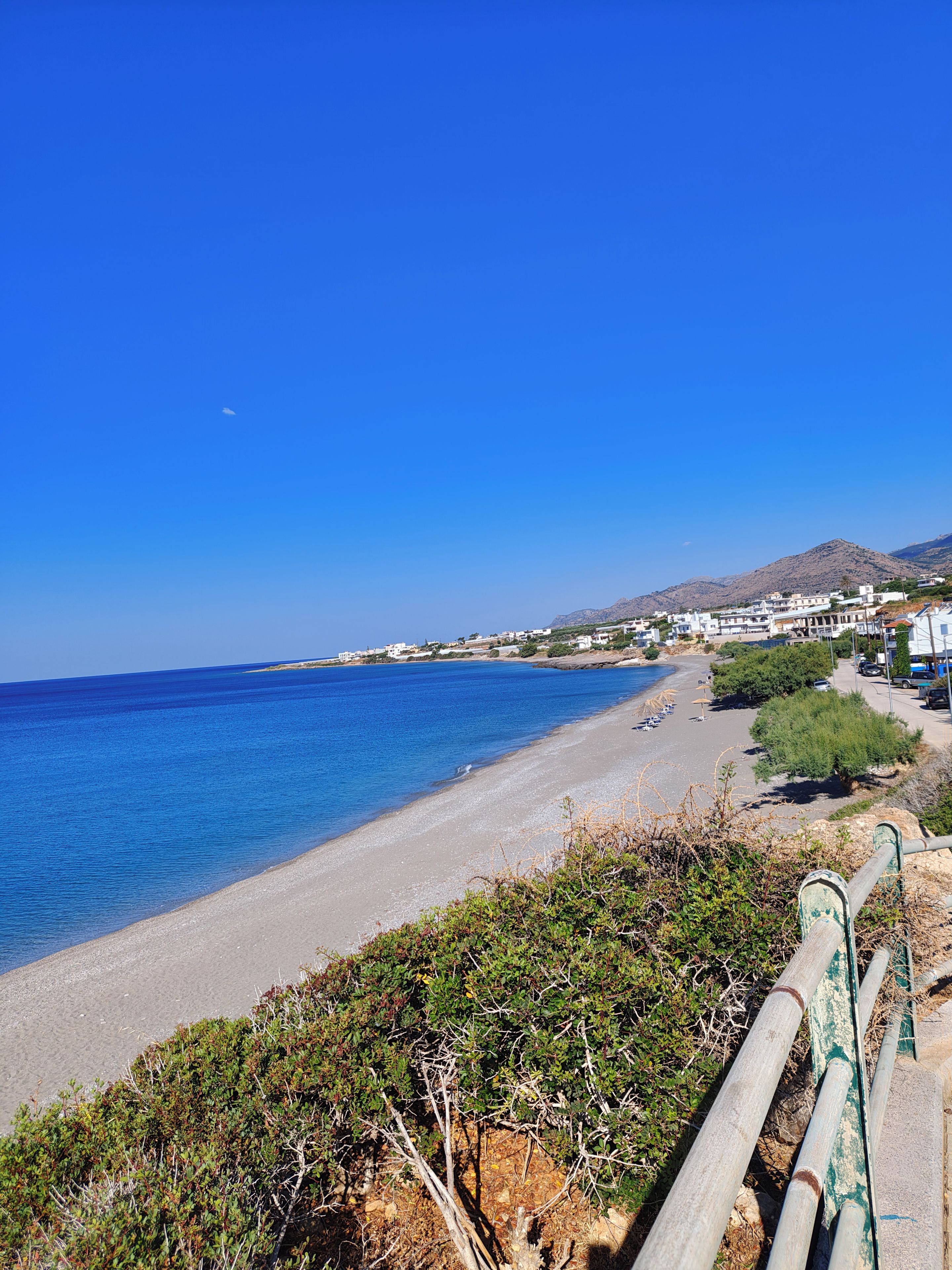 A coastal view with a clear sky, a sandy beach, and buildings in the distance.