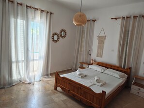 A bedroom with a wooden bed, sheer curtains, a woven light fixture, and wall decor. - Spacious Apartment Garden View Partial Seaview (Makrygialos)