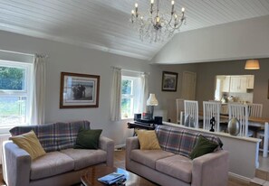 A living room with two sofas, a coffee table, and a chandelier. There is a kitchen area visible through an open doorway. - River Cottage (Clifden)