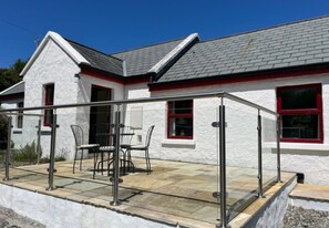A white building with a grey roof, a covered patio with chairs, and a glass railing. - River Cottage (Clifden)